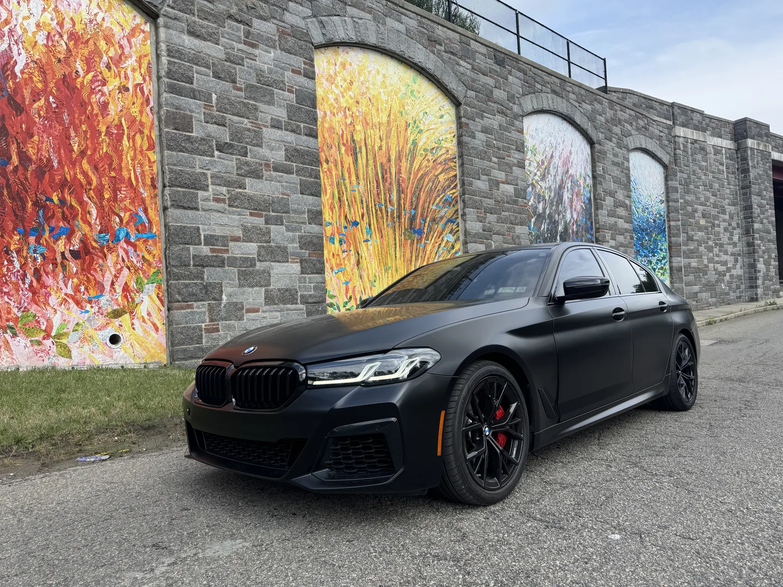 BMW 5 Series in full satin black wrap parked beside a stone wall with painted murals